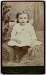 Child in white dress sitting on hay, front