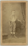 Boy standing with hand rested over small table wearing a cape and long style boots, hair combed rear with curls at end, front by A. Parker